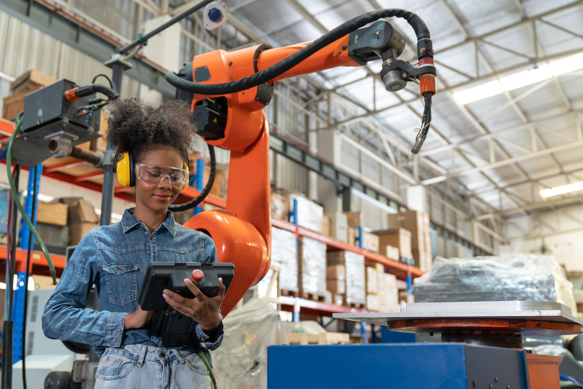 A young African American female engineer operating an industrial robot in a warehouse, holding a control pad and wearing safety glasses and hearing protection.
